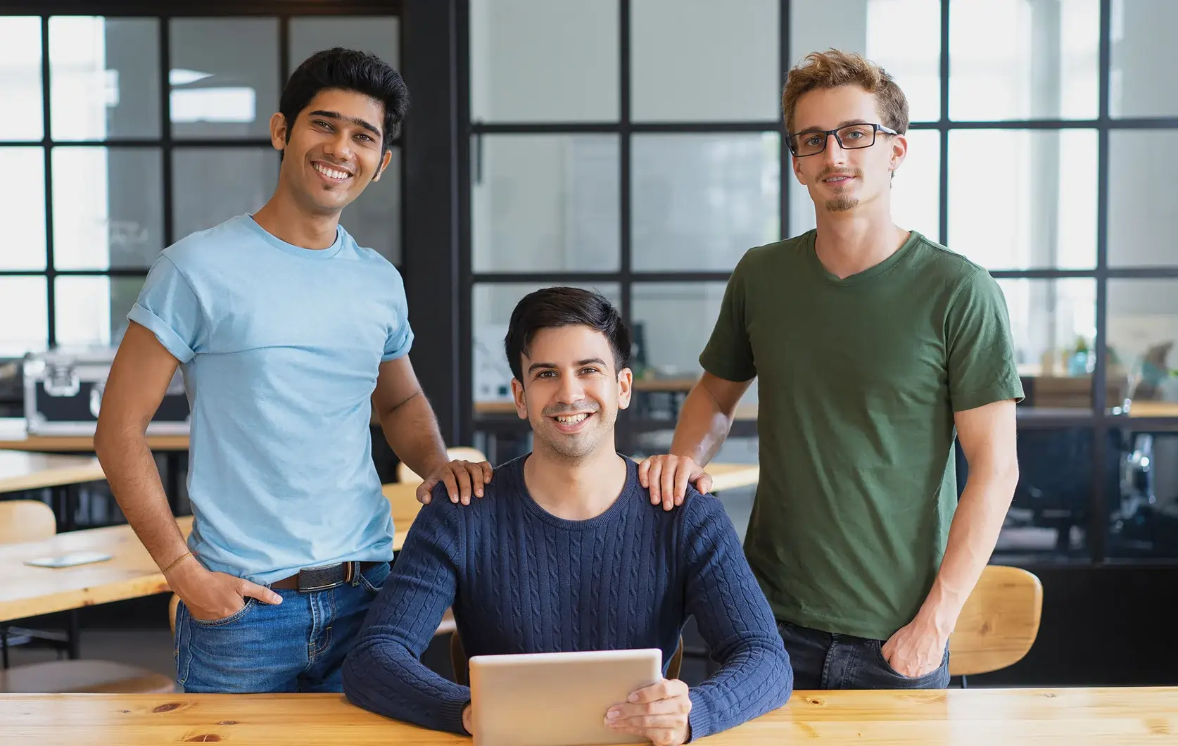 Three men in casual clothing pose together in a modern office. Two stand while one sits at a table holding a tablet. They are smiling and looking at the camera.