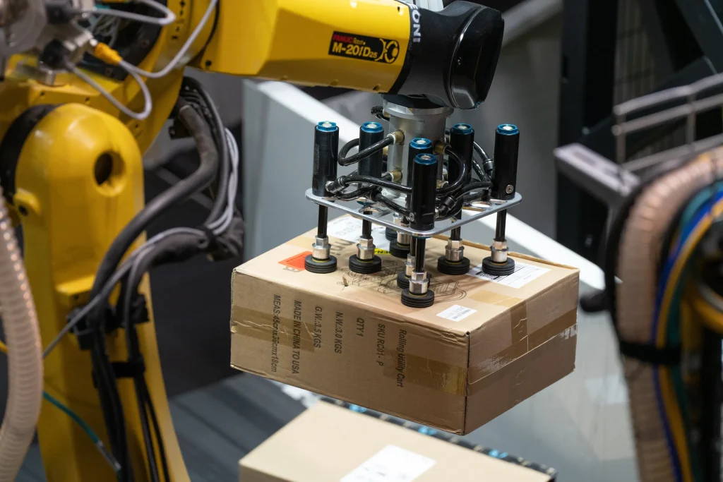 A yellow industrial robotic arm uses a suction gripper to lift a cardboard box in a warehouse, showing automation in logistics